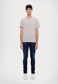 T-shirt grigio a maniche corte, jeans slim-fit blu scuro, sneaker bianche, cappellino nero con testo, in piedi su uno sfondo bianco semplice.
