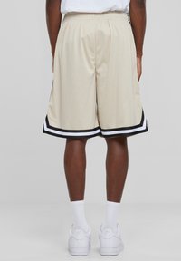Beige mesh basketball shorts med sorte og hvide striber, elastisk talje, knælangt design og sidesplit-detaljer.