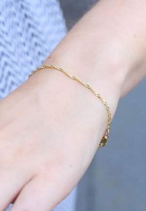 Gouden armband om een slanke pols, met een gedraaid ontwerp en een sluiting met een klein accentstuk. Glad van textuur. Minimalistische stijl.