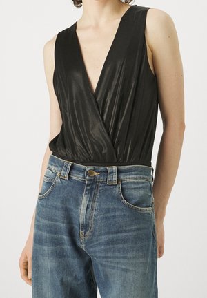 Persoon die een mouwloze zwarte wikkel-top draagt, ingestopt in een hoog uitgesneden blauwe spijkerbroek met knoop en voorzakken.