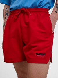 Rote Sportshorts mit Kordelzugbund und „hummel SPORTSWEAR“-Logo, getragen von einer tätowierten Person mit Hand in der Tasche.