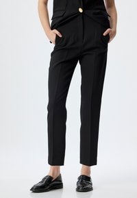 Pantalons noirs ajustés avec une coupe droite, dotés de poches latérales et de plis subtils, fabriqués en tissu lisse. Portés avec des mocassins noirs.