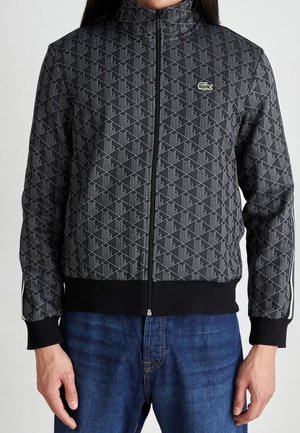 Personne portant une veste noire zippée à motif avec un logo de crocodile vert sur la poitrine et un jean en denim bleu.
