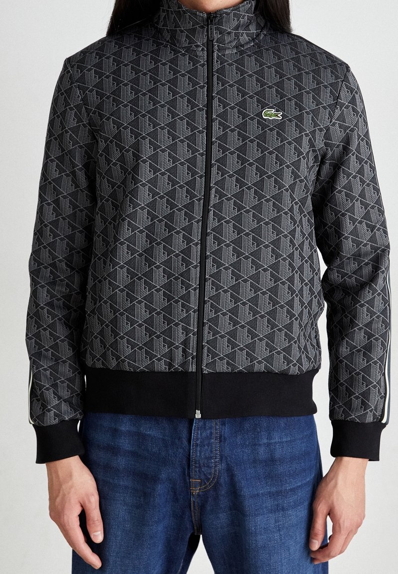 Personne portant une veste noire zippée à motif avec un logo de crocodile vert sur la poitrine et un jean en denim bleu.