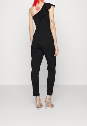 Mono negro con un hombro y detalle de volantes, piernas ajustadas, tejido texturizado y diseño de espalda acentuado. Combinado con tacones altos plateados.