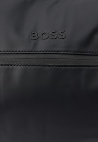 Zbliżenie na czarną tkaninę z wytłoczonym logo "BOSS" nad poziomym szwem zamka błyskawicznego.