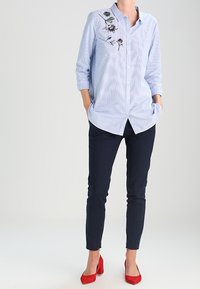 Camicia azzurra a righe con dettagli floreali ricamati, abbinata a pantaloni slim-fit blu navy e tacchi rossi.