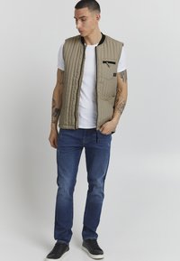 Beige quiltet vest med glidelåsforkant og lomme, svart krage, kombinert med blå jeans og svarte joggesko. Moderne, avslappet design.