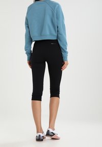 Felpa blu corta con maniche lunghe, leggings neri fino al ginocchio e scarpe da ginnastica bianche con dettagli rosa, in piedi contro uno sfondo neutro.