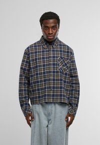 Férfi kék, zöld és barna kockás ingben, amelynek dőlt mellzsebe van, világoskék, laza szabású farmernadrággal párosítva, fehér háttér előtt.