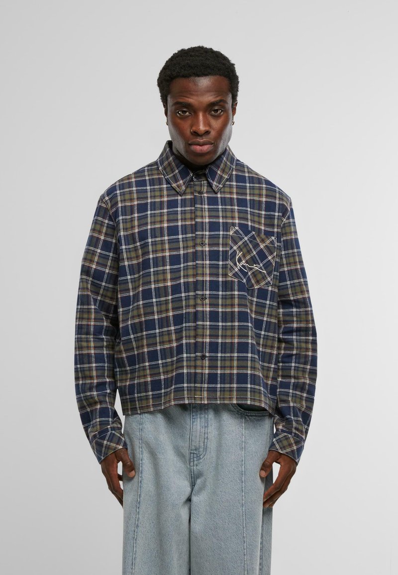 Férfi kék, zöld és barna kockás ingben, amelynek dőlt mellzsebe van, világoskék, laza szabású farmernadrággal párosítva, fehér háttér előtt.