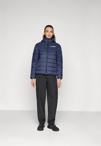 Marineblaue Steppjacke mit Kapuze, ausgestattet mit horizontalen Nähten und einem Frontreißverschluss. Kombiniert mit schwarzen Cargohosen und dunklen Sneakers.