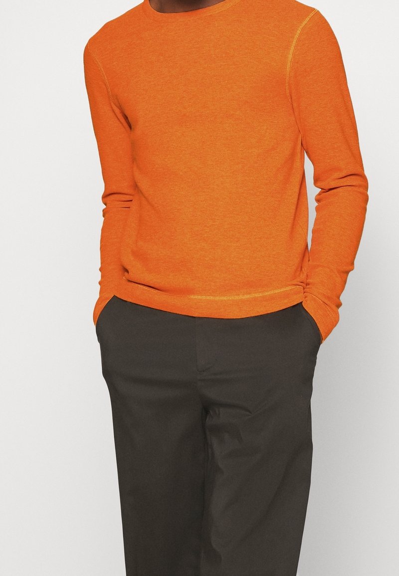 Personne portant un pull à manches longues orange vif et un pantalon gris foncé, debout les mains dans les poches devant un fond uni.