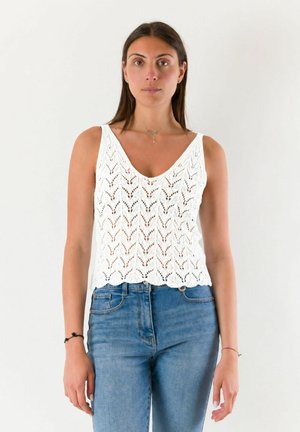 Giovane donna che indossa un top bianco in maglia senza maniche e jeans blu a vita alta, in piedi davanti a uno sfondo chiaro e semplice.