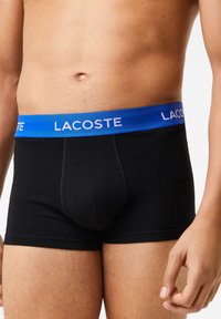 Boxer-briefs en coton noir avec une ceinture élastique bleue arborant le logo "LACOSTE" en blanc. Détails cousus sur le tissu.