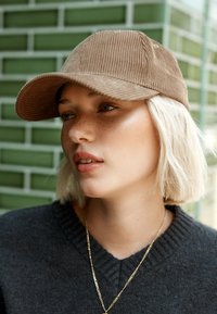 Casquette en velours côtelé marron avec une visière incurvée, présentant des rainures verticales, associée à un pull foncé et une fine chaîne en or. Fond texturé vert.