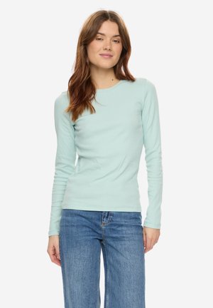 Vrouw draagt een lichtblauw nauwsluitend shirt met lange mouwen en een blauwe spijkerbroek, staat met een neutrale uitdrukking tegen een effen achtergrond.