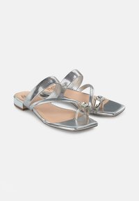 Zilveren metallic platte sandalen met dubbele kruislings gestructureerde banden, vierkante teenontwerp en een decoratieve edelsteenaccent aan de voorkant. Glad textuur.