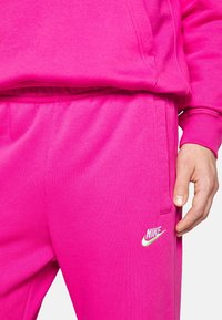 Osoba v jasně růžových teplákových kalhotách Nike a odpovídajícím mikině, ukazuje pravou ruku a logo Nike na kapse kalhot.