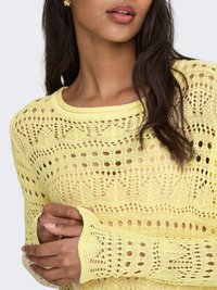 Vrouw met een gele gebreide trui met kantpatroon, die met één hand de mouw vasthoudt, gouden oorbellen draagt en lang donker haar heeft.