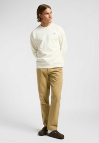 Sweatshirt crème clair avec un petit logo sur la poitrine, associé à un pantalon beige et des sabots marron. Look simple et décontracté.