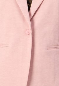 Blazer rose clair en tissu extensible avec col cranté, fermeture à un bouton et deux poches avant. Texture lisse, design décontracté.