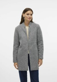 Grau-wolliger Mantel mit strukturierter Oberfläche, offenem Frontbereich und Stehkragen, kombiniert mit einem cremefarbenen Pullover und dunklen Jeans.