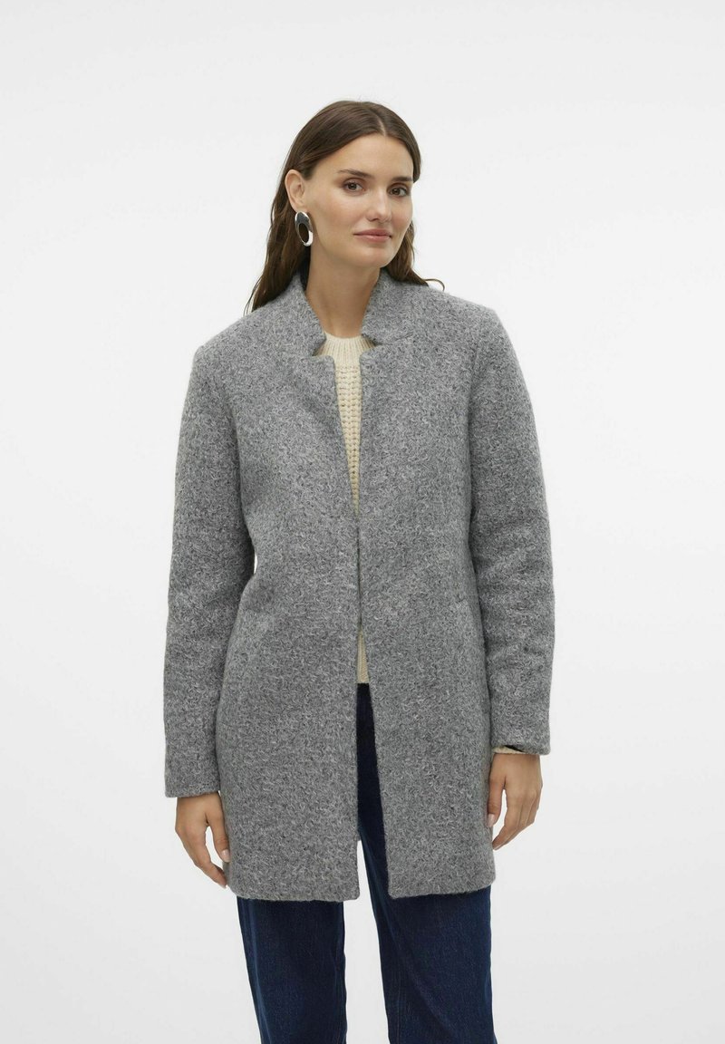 Grau-wolliger Mantel mit strukturierter Oberfläche, offenem Frontbereich und Stehkragen, kombiniert mit einem cremefarbenen Pullover und dunklen Jeans.