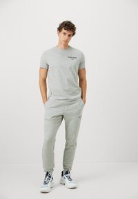 Harmaa puuvillapaita, jossa on lyhyet hihat ja musta logo rinnassa, yhdistettynä harmaisiin jogging-housuihin, joissa on joustava vyötärö ja logokoristeluja.