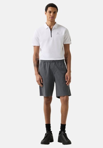 Young man standing, wearing white short-sleeve polo, gray drawstring shorts, black socks, and black sneakers on a plain background.