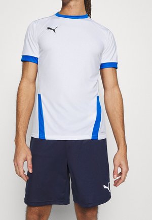 Man draagt een wit sportshirt met blauwe bies en donkerblauwe shorts met Puma-logo, staand tegen een effen achtergrond.