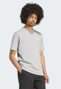 Šedé bavlněné tričko s kulatým výstřihem, krátkými rukávy a bílým logem Adidas na levé hrudi, spolu s černými kalhotami.