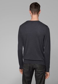 Muž otočený zády, na sobě má tmavě šedý svetr s dlouhým rukávem a černé džíny na jednobarevném šedém pozadí.