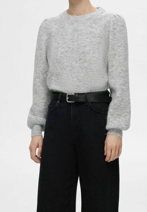 Persona vistiendo un suéter de punto gris claro con mangas abullonadas, metido dentro de pantalones negros de cintura alta asegurados con un cinturón negro.