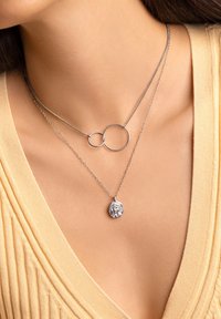 Collares de plata en capas, uno con círculos entrelazados y el otro con un colgante texturizado que representa una cara, llevados sobre una blusa beige.