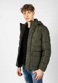 Gevoerde groene jas met een rits en drukknoopsluiting, voorzien van een capuchon, twee voorkanten zakken en ribgebreide manchetten. Draagt over een zwart T-shirt.