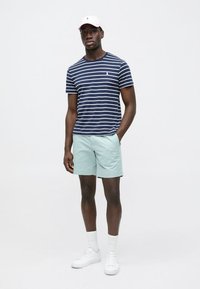 T-shirt azul marinho às riscas brancas, calções verdes claros, ténis brancos e boné rosa claro. Modelo de pé com as mãos nos bolsos.