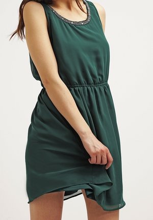 Kobieta w bezrękawną ciemnozieloną sukienkę z koralikowym dekoltem, trzymająca jedną ręką brzeg sukienki.