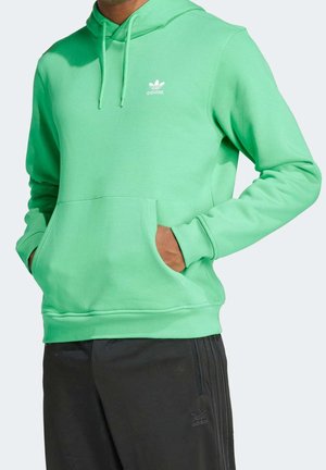 Person trägt einen leuchtend grünen Adidas-Hoodie mit Kängurutasche und schwarze Adidas-Trainingshose mit Logo am linken Oberschenkel.