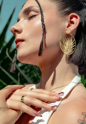 Large circular gold earrings with a leaf design, worn by a person with side-braided hair, displaying a close-up profile against a blue background.