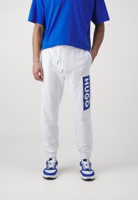 Weiße Jogginghose mit blauem Logo "HUGO" an der Seite, kombiniert mit einem blauen Oversized-T-Shirt und blau-weißen Sneakers. Lässiges Design.