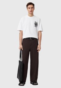 Λευκό βαμβακερό t-shirt με κοντά μανίκια και γραφικό σχέδιο στο αριστερό στήθος, συνδυασμένο με καφέ παντελόνες με φαρδιά πόδια και μαύρη τσάντα tote.