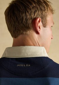Marineblau gestreiftes Rugby-Shirt mit einem cremfarbenen Kragen. Der Ausschnitt zeigt ein goldenes "JOULES"-Logo. Der Stoff wirkt strukturiert.