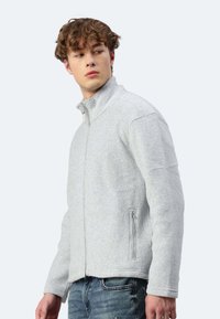 Giacca in pile grigia chiara con colletto alto, zip frontale e tasche laterali. Presenta una consistenza morbida e un design aderente.