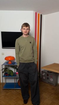 Νέος άνδρας με κοντά καστανά μαλλιά, ντυμένος με πράσινο φούτερ και μαύρο παντελόνι, στέκεται σε δωμάτιο με πορτοκαλί λάμπα, τηλεόραση και πολύχρωμες ρίγες στον τοίχο.