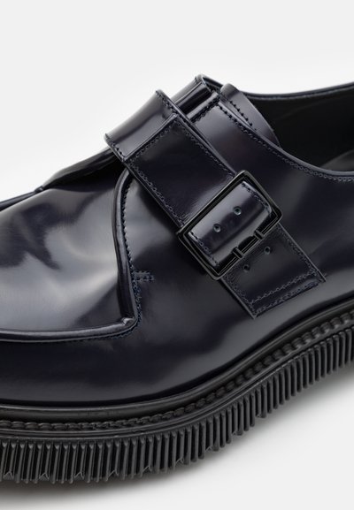 Pantofii din piele patenta de culoare bleumarin au un vârf pătrat, detalii cu curea cu cataramă și o talpă din cauciuc texturat negru. Prezintă accente cusute de-a lungul marginilor.