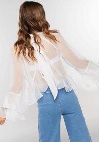 Blusa blanca transparente con mangas fluídas y un lazo en la parte posterior, combinada con jeans de tiro alto azules. Textura suave y elementos de diseño en capas.