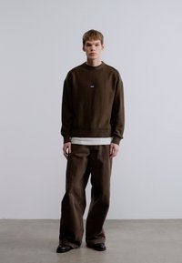 Pull marron foncé avec un col côtelé, superposé sur une chemise blanche, assorti à un pantalon marron à jambes larges. Texture lisse, coupe décontractée.