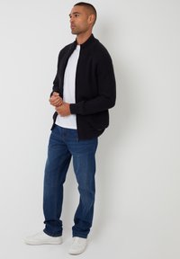 Gestrickter schwarzer Reißverschluss-Pullover mit hohem Kragen, getragen über einem weißen T-Shirt, kombiniert mit blauen Jeans und weißen Sneakern.