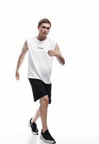 Biała bezrękawnikowa koszula z napisem "UNSTOPPABLE", czarne szorty sportowe i czarne sneakersy, prezentujące szkieletowy design oraz casualowy styl.